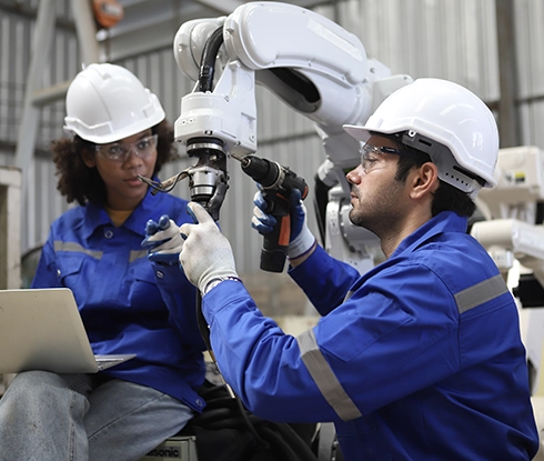 Two workers wearing safety equipment work on repairing a robotic arm.