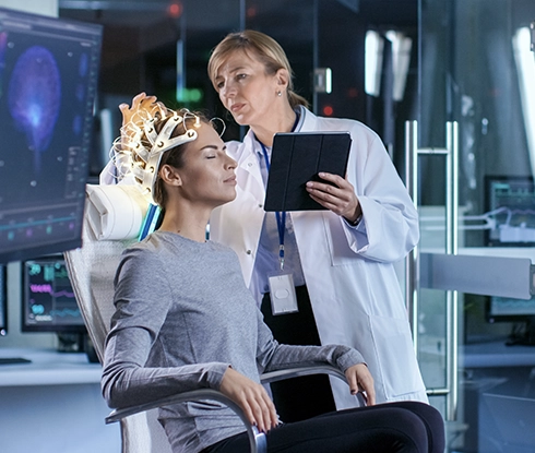 A woman sitting in a chair with neural sensors attached to her head is inspected by a doctor. The doctor has a tablet in her hand.