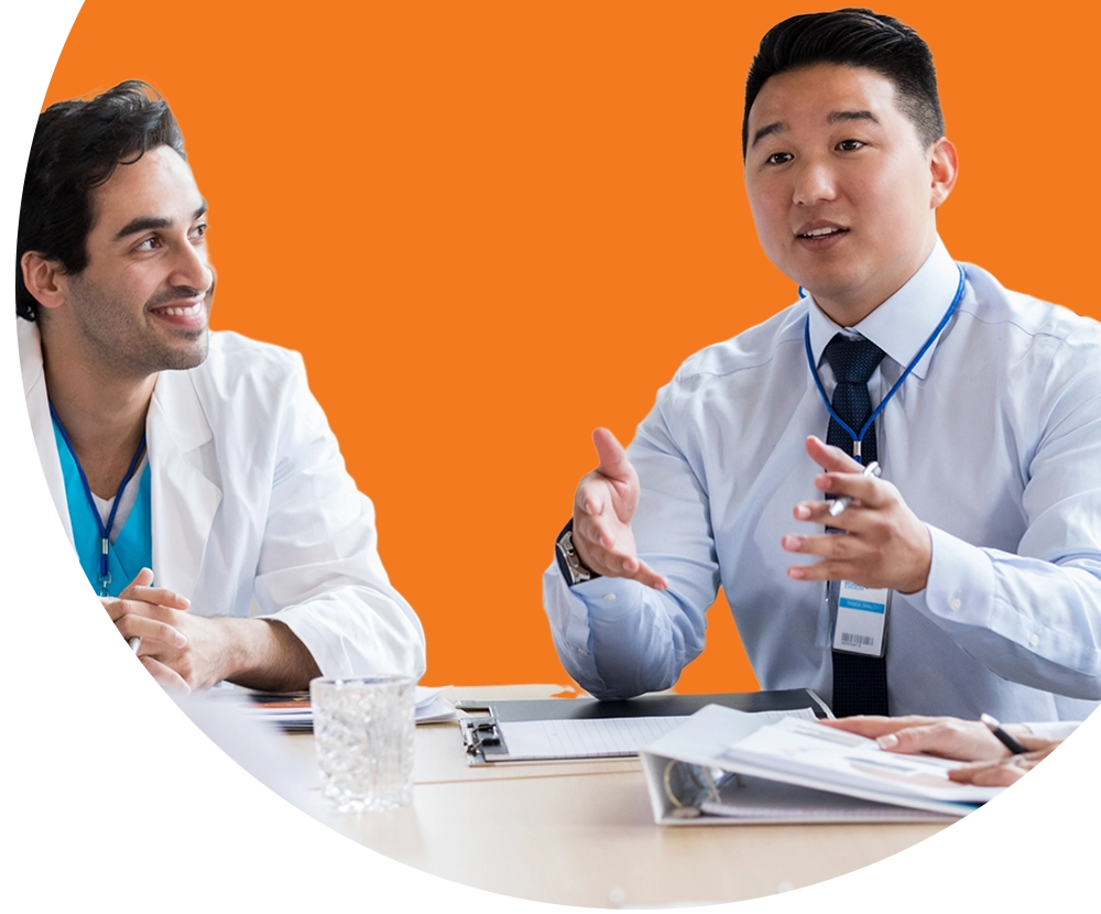 Two medical professionals sitting at a table. One is wearing a lab coat and the other business attire.