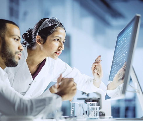 Two people wearing lab coats look at a computer monitor